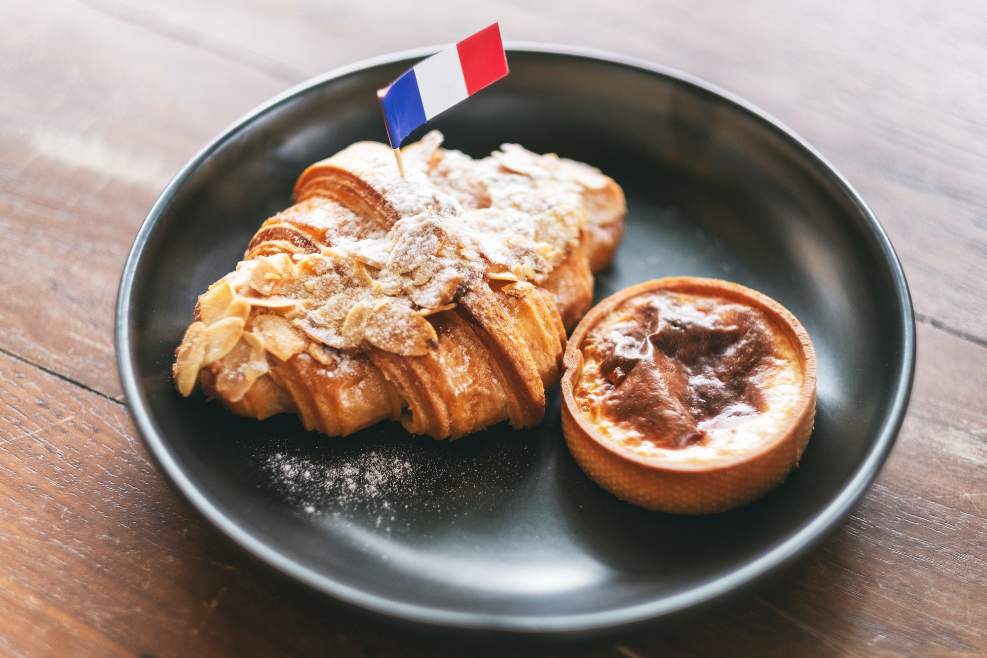 A piece of croissant and tart with a flag of France in a black plate on wooden table in cafe