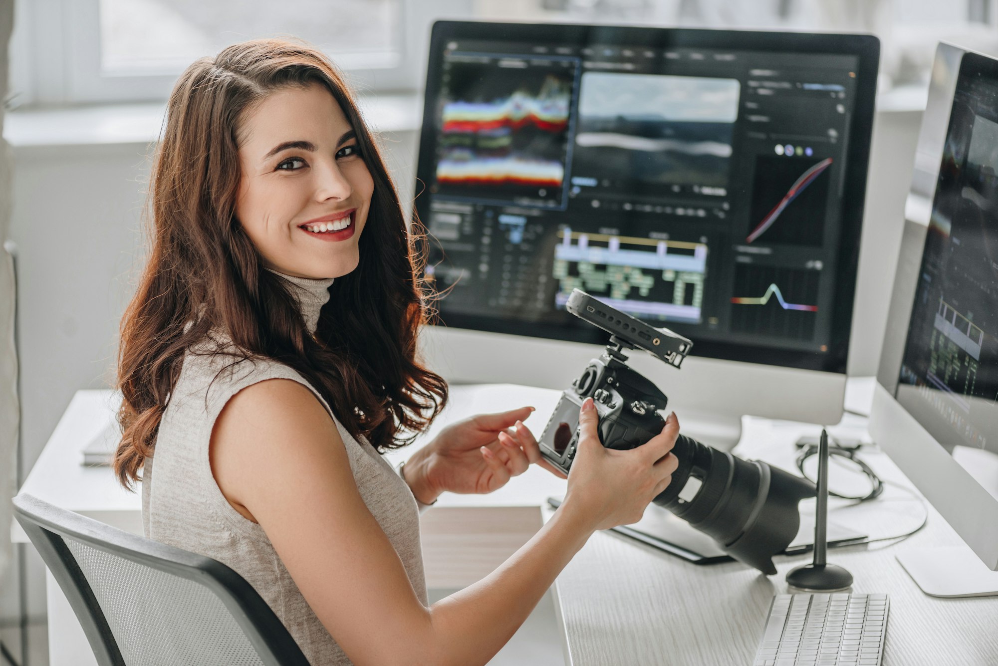 cheerful art editor holding digital camera near table with computer monitors