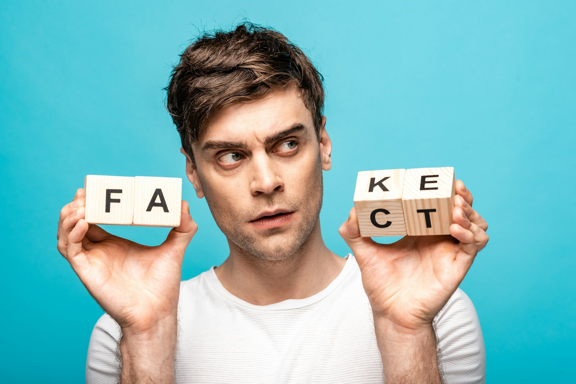 thoughtful young man looking away while holding wooden cubes with fake fact lettering isolated on