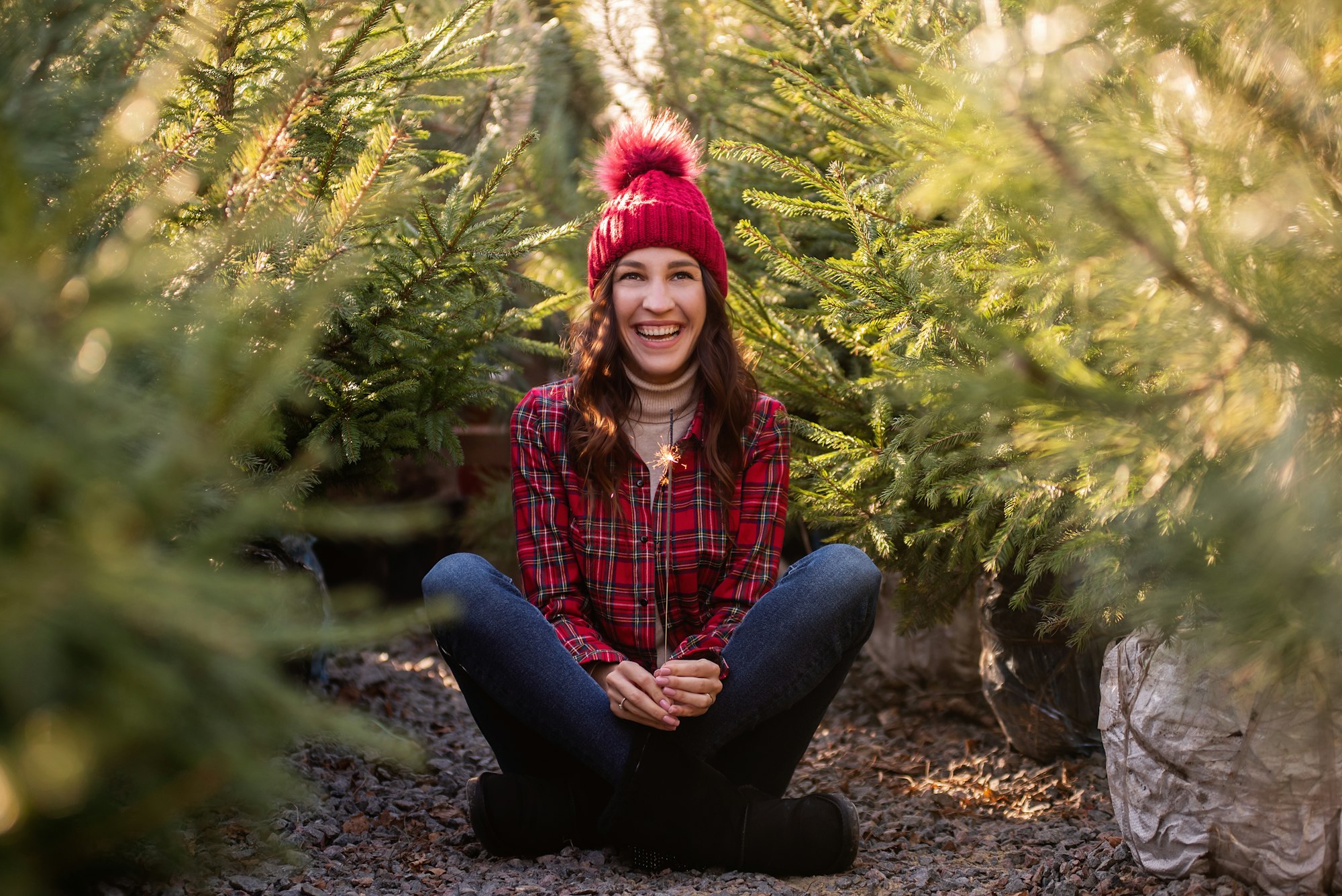 Young woman with sparkler in hand wearing red checkered shirt, hat among green Christmas tree market